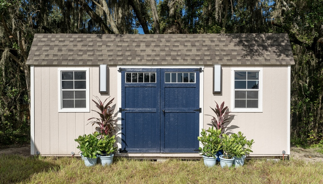 Front facing view of a cream colored shed with blue double doors with nice entry way plants on wither side of the doors.